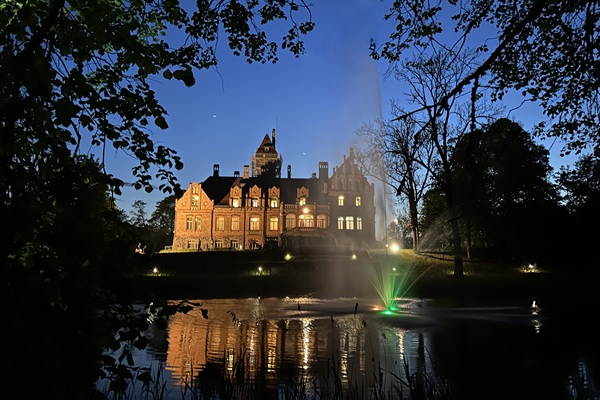 Izgaismota LVM Jaunmoku pils naktī ar dīķi un strūklaku priekšplānā